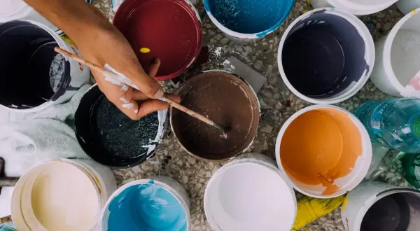 Ręka z pędzlem miesza farby w wiaderkach. Widać różne kolory, co może sugerować wady farby ceramicznej, np. nierównomierne krycie.