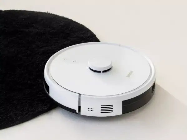 Biały robot sprzątający obok czarnego dywanika. Jego moc ssania odkurzacza poradzi sobie z każdym brudem.